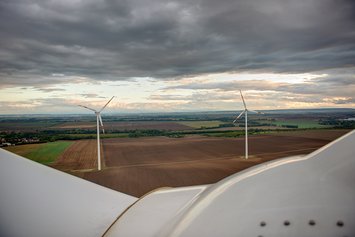 Luftaufnahme einer Windkraftanlage. Im Hintergrund sind bewirtschaftete Felder mit zwei Windkraftanlagen zu sehen.