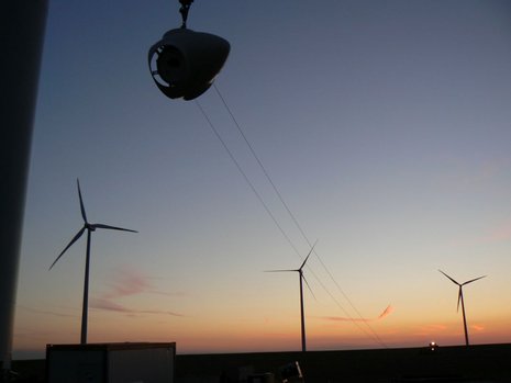 Eine Rotornabe wird bei Sonnenuntergang in die Luft gehoben. Im Hintergrund sind drei Windkraftanlagen zu sehen.