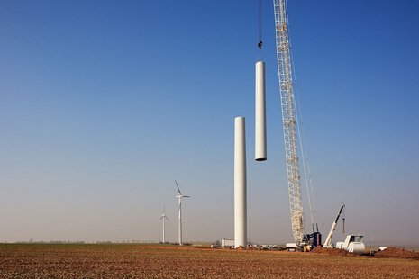 Fernaufnahme der Mastmontage einer Windkraftanlage mit einem weißen Kran. In der Ferne sind zwei Windkraftanlagen zu sehen.
