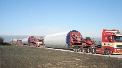 3 rote LKWs transportieren Teile eines Windradmastes über eine Straße mitten auf einem Feld.