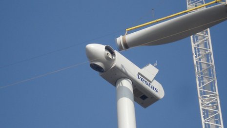 Aufnahme von unten bei der Montage eines Windradblattes am Rotor mit einem weißen Kran.