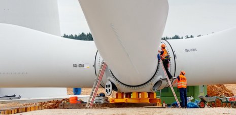 Montage der Rotorblätter an der Windkraftanlage. Zwei Arbeiter koordinieren den Vorgang.
