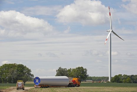 Fernaufnahme eines Feldes mit einer Windkraftanlage und einem Transportfahrzeug in der Mitte. Bäume stehen daneben.