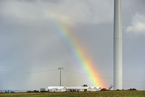 Regenbogen mit einer Windkraftanlage daneben und weißen Zelten auf dem Boden.