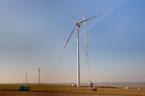 Ein Feld mit einer Windkraftanlage in der Mitte und einem gelben Kran daneben. Im Hintergrund sind zwei weitere Windkraftanlagen und ein Bulldozer zu sehen.